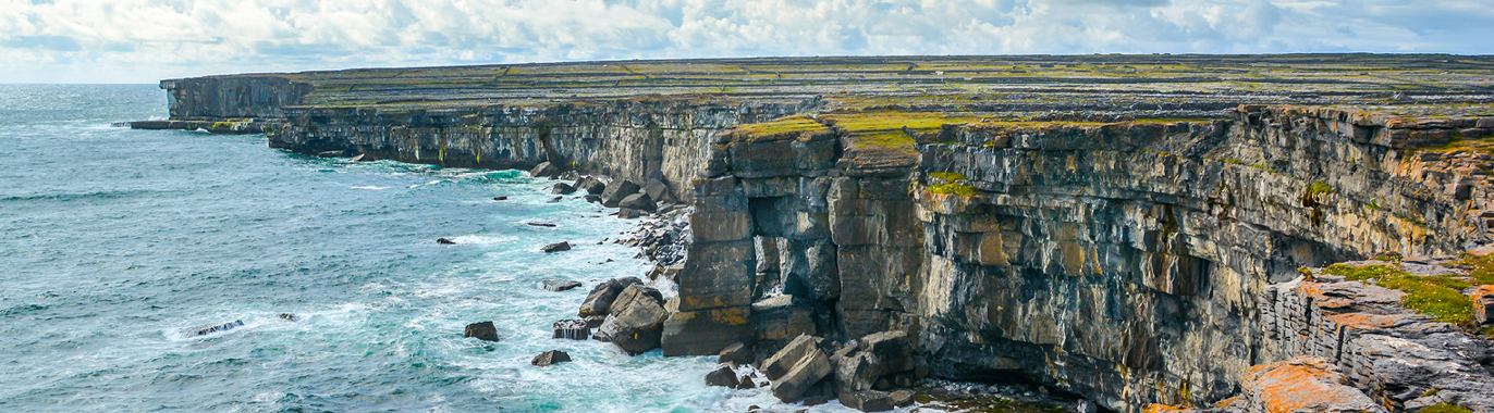 Rocky sea cliffs with layered stone formations overlook the ocean under a partly cloudy sky, with waves crashing at the base and sparse vegetation on top—an iconic scene from the Emerald Isle countryside.