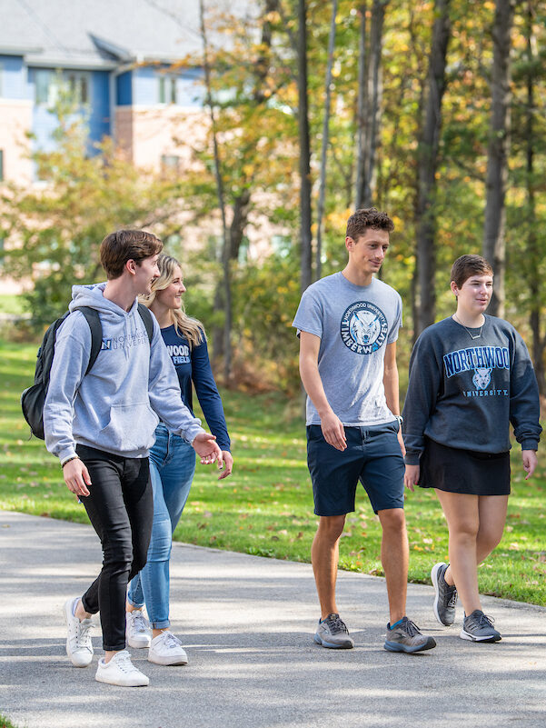 Five students walk together on a campus path lined with trees, wearing casual clothing and university-branded apparel—capturing the spirit of a Visit Campus experience.