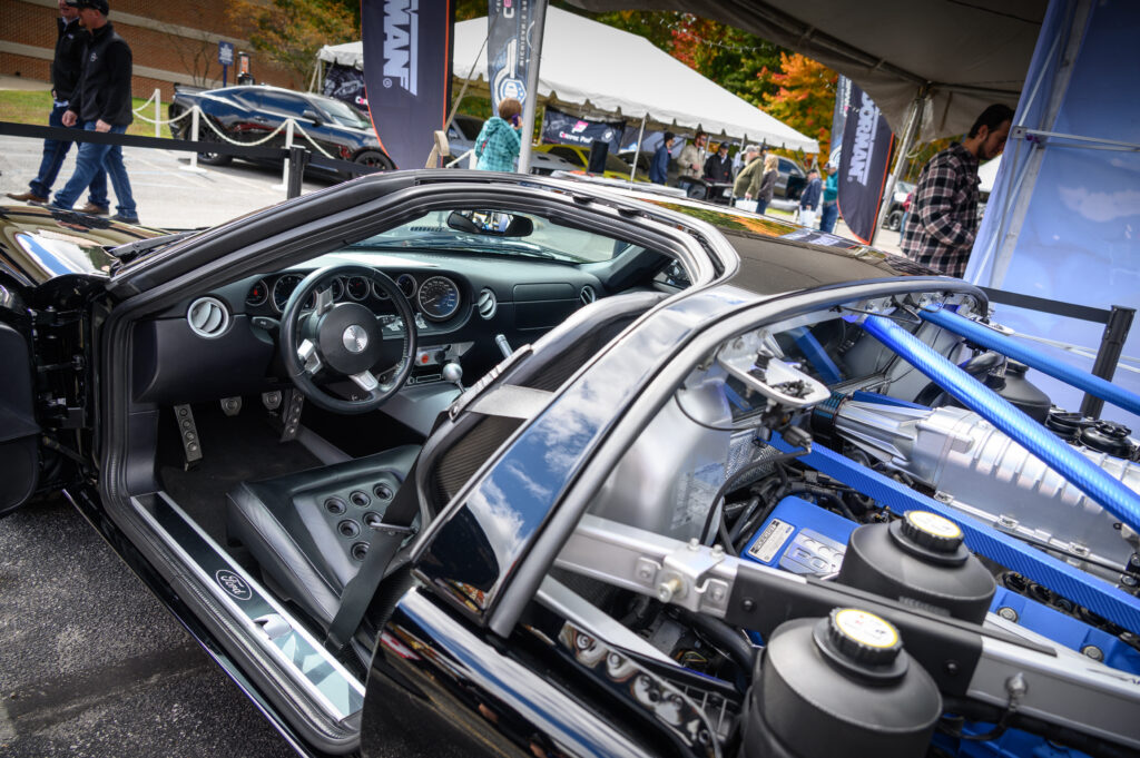 Close-up view of a sports car interior and engine bay at an outdoor car show, highlighting Automotive Aftermarket Management features, with people and tents visible in the background.