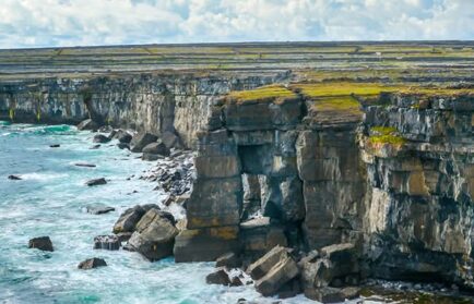 Rocky sea cliffs with green grassy tops rise above crashing waves, under a cloudy sky—an iconic scene for your 2026 Trip to the Emerald Isle.