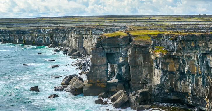 Rocky sea cliffs with green grassy tops rise above crashing waves, under a cloudy sky—an iconic scene for your 2026 Trip to the Emerald Isle.