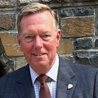 A man in a suit and tie stands in front of a stone wall, smiling slightly at the camera, embodying Leadership Insights with his confident and approachable demeanor.