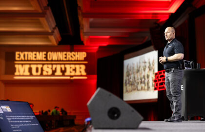 A speaker stands on stage giving a presentation at an "Extreme Ownership Muster" event, with a projected image and audience visible in the background.