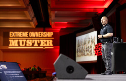A speaker stands on stage giving a presentation at an "Extreme Ownership Muster" event, with a projected image and audience visible in the background.