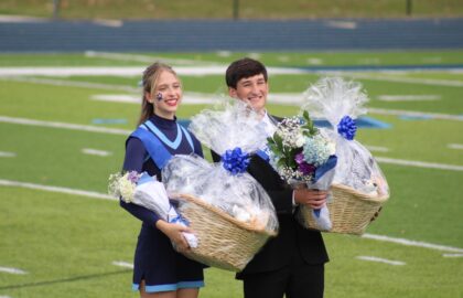 A cheerleader and a young man in a suit stand on a football field, each holding a large gift basket and bouquet wrapped in cellophane.