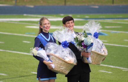 A cheerleader and a young man in a suit stand on a football field, each holding a large gift basket and bouquet wrapped in cellophane.
