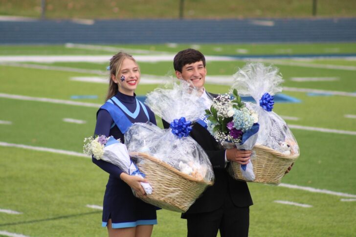 A cheerleader and a young man in a suit stand on a football field, each holding a large gift basket and bouquet wrapped in cellophane.