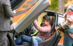 A man sits inside a sports car with gullwing doors open, smiling as others stand nearby at the Auto Show; the car displays a "#McLarenRally" decal, promising elevated experiences from Sept. 19-21.