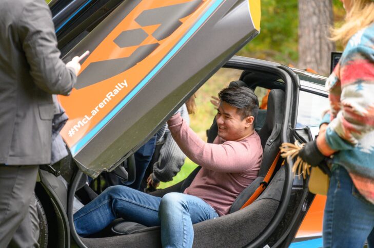 A man sits inside a sports car with gullwing doors open, smiling as others stand nearby at the Auto Show; the car displays a "#McLarenRally" decal, promising elevated experiences from Sept. 19-21.