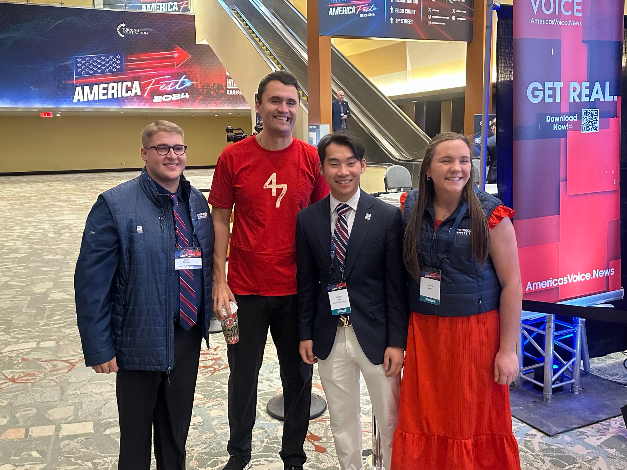 Four people stand together indoors at a Turning Point USA Northwood event, with America-themed banners and signage in the background. The group poses and smiles for the photo with Charlie Kirk.