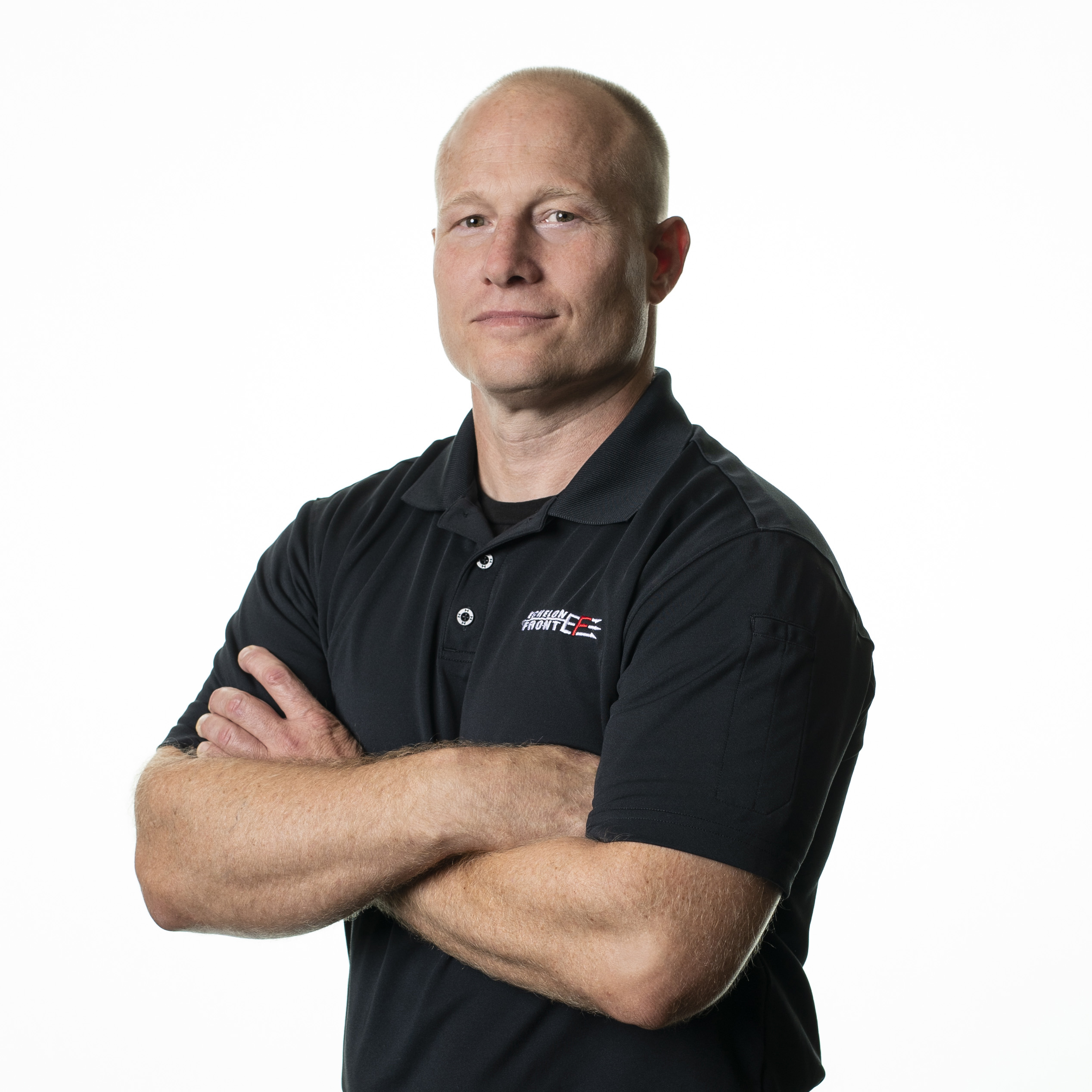 A man with a shaved head wearing a black collared shirt stands with his arms crossed against a plain white background, exuding confidence and offering Leadership Insights through his poised demeanor.