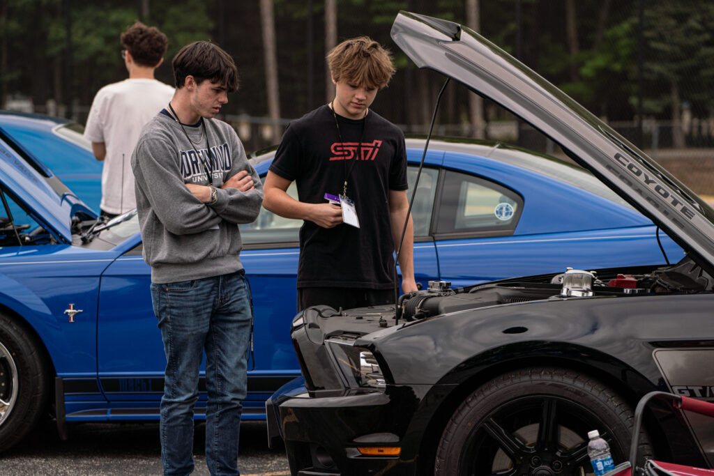 Two young men stand next to a black car with its hood open, examining the engine bay—perhaps discussing Automotive Aftermarket Management—while a blue car is parked nearby in an outdoor setting.