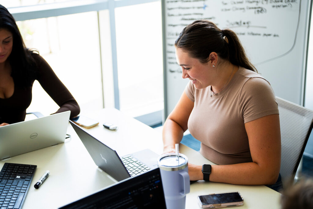 A woman sits at a table working on a laptop, possibly accessing SAP software, with a tumbler and phone nearby. Another person is partially visible next to her. A whiteboard is seen in the background.