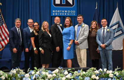 Nine people stand in a row, dressed in formal attire, in front of a Northwood University backdrop with floral arrangements, celebrating the Outstanding Alumni of 2025.
