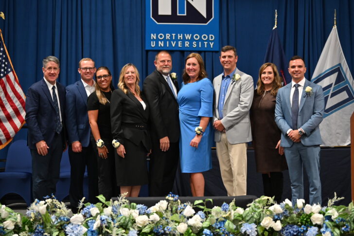 Nine people stand in a row, dressed in formal attire, in front of a Northwood University backdrop with floral arrangements, celebrating the Outstanding Alumni of 2025.