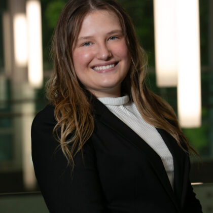 A woman with long, wavy hair wearing a black blazer and white top smiles at the camera in an indoor setting with blurred lights in the background.