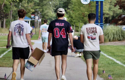 Three young men walk down a campus sidewalk, two wearing Kappa Sigma shirts, one holding a box. Small American flags line the path. Trees and a campus sign are visible.