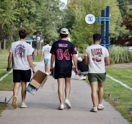 Three young men walk down a campus sidewalk, two wearing Kappa Sigma shirts, one holding a box. Small American flags line the path. Trees and a campus sign are visible.