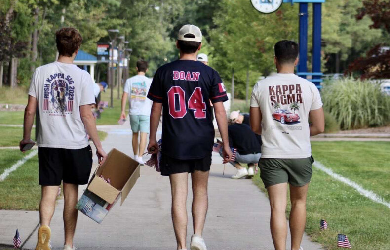Three young men walk down a campus sidewalk, two wearing Kappa Sigma shirts, one holding a box. Small American flags line the path. Trees and a campus sign are visible.