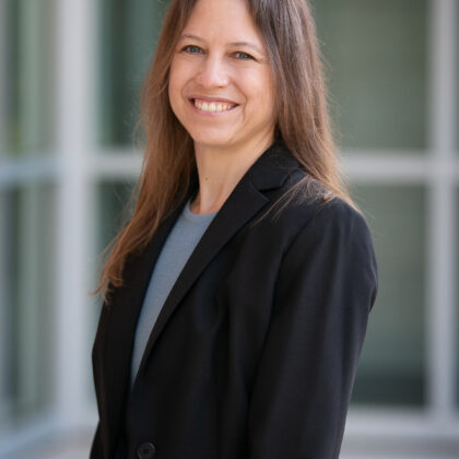 A woman with long brown hair wearing a black blazer and blue top stands indoors, smiling at the camera, with blurred glass windows in the background.