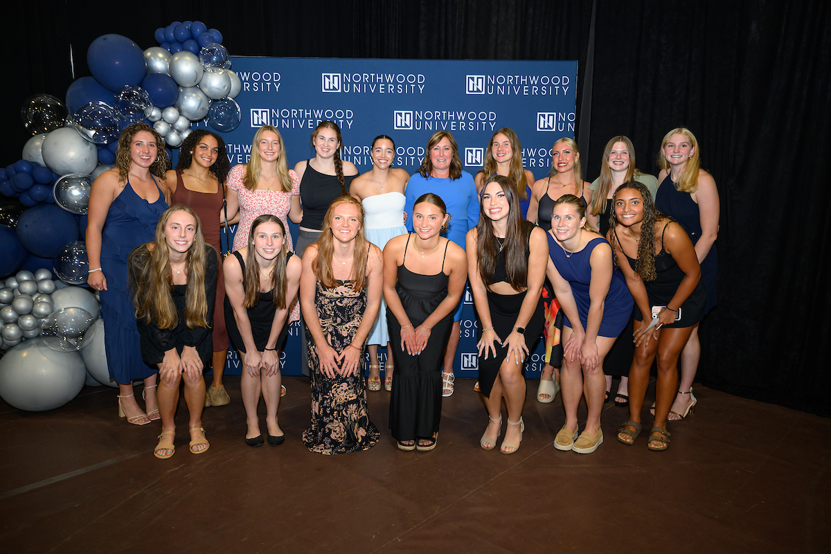 A group of sixteen young women pose together in front of a Northwood University backdrop and blue balloon decorations at an indoor event celebrating Northwood's Outstanding Alumni Awards.