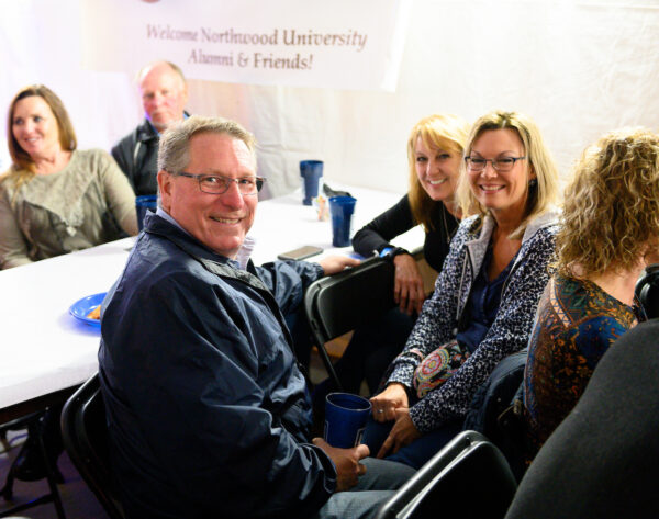 A group of adults sit at a table, smiling at the camera during a Northwood University alumni and friends event, enjoying conversations about Alumni Services and Benefits.