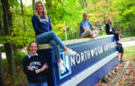 Five students pose and smile on and around a large Northwood University sign outdoors, surrounded by green trees and rocks, celebrating support from the Gerstacker Foundation for middle-income students.
