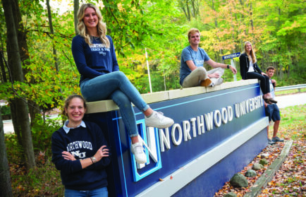 Five students pose and smile on and around a large Northwood University sign outdoors, surrounded by green trees and rocks, celebrating support from the Gerstacker Foundation for middle-income students.