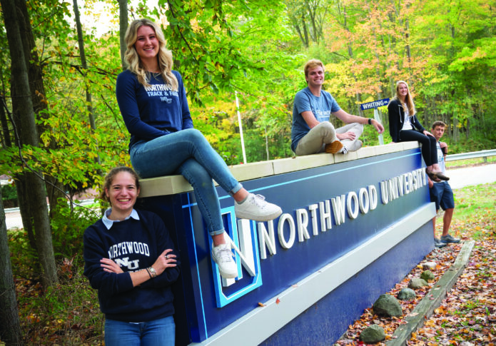 Five students pose and smile on and around a large Northwood University sign outdoors, surrounded by green trees and rocks, celebrating support from the Gerstacker Foundation for middle-income students.