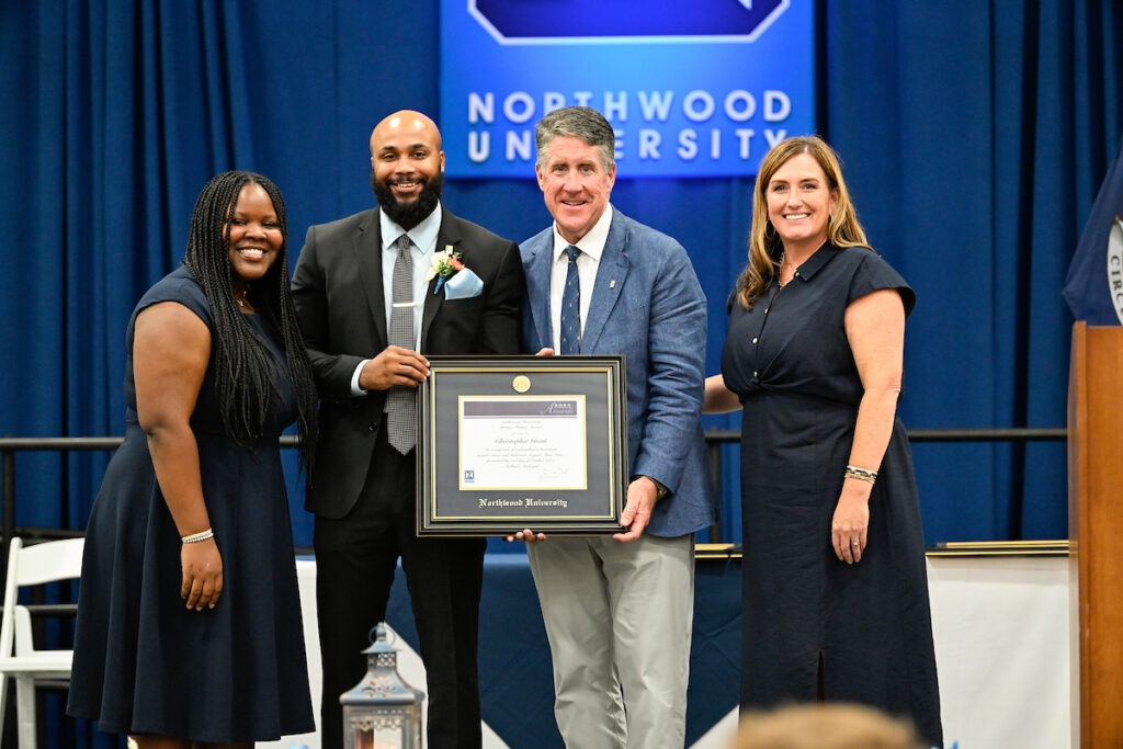 Four people stand on a stage at Northwood University, with one man holding a framed certificate, posing for a group photo during Northwood's Outstanding Alumni Awards.