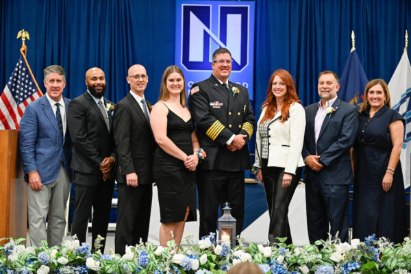Eight people in formal attire stand in a row on a stage with blue curtains, floral arrangements, and flags, with a uniformed officer at the center—capturing the spirit of annual alumni events and networking.