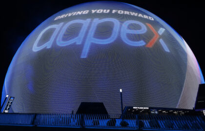 At night, a large spherical display lights up with "DRIVING YOU FORWARD," "aapex," and a nod to AAPEX 2025 in bold blue and red letters.
