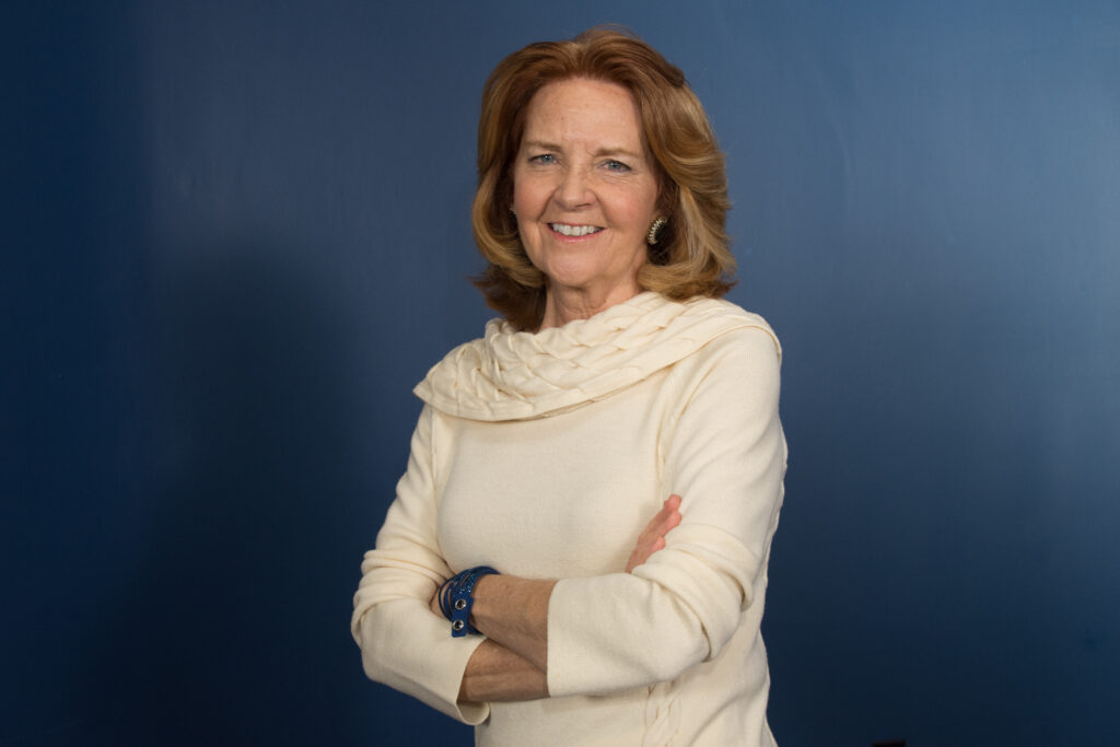 A woman with shoulder-length reddish hair stands against a blue background, smiling with her arms crossed and wearing a cream-colored sweater—perfectly capturing the spirit of the Northwood's Distinguished Women Awards.