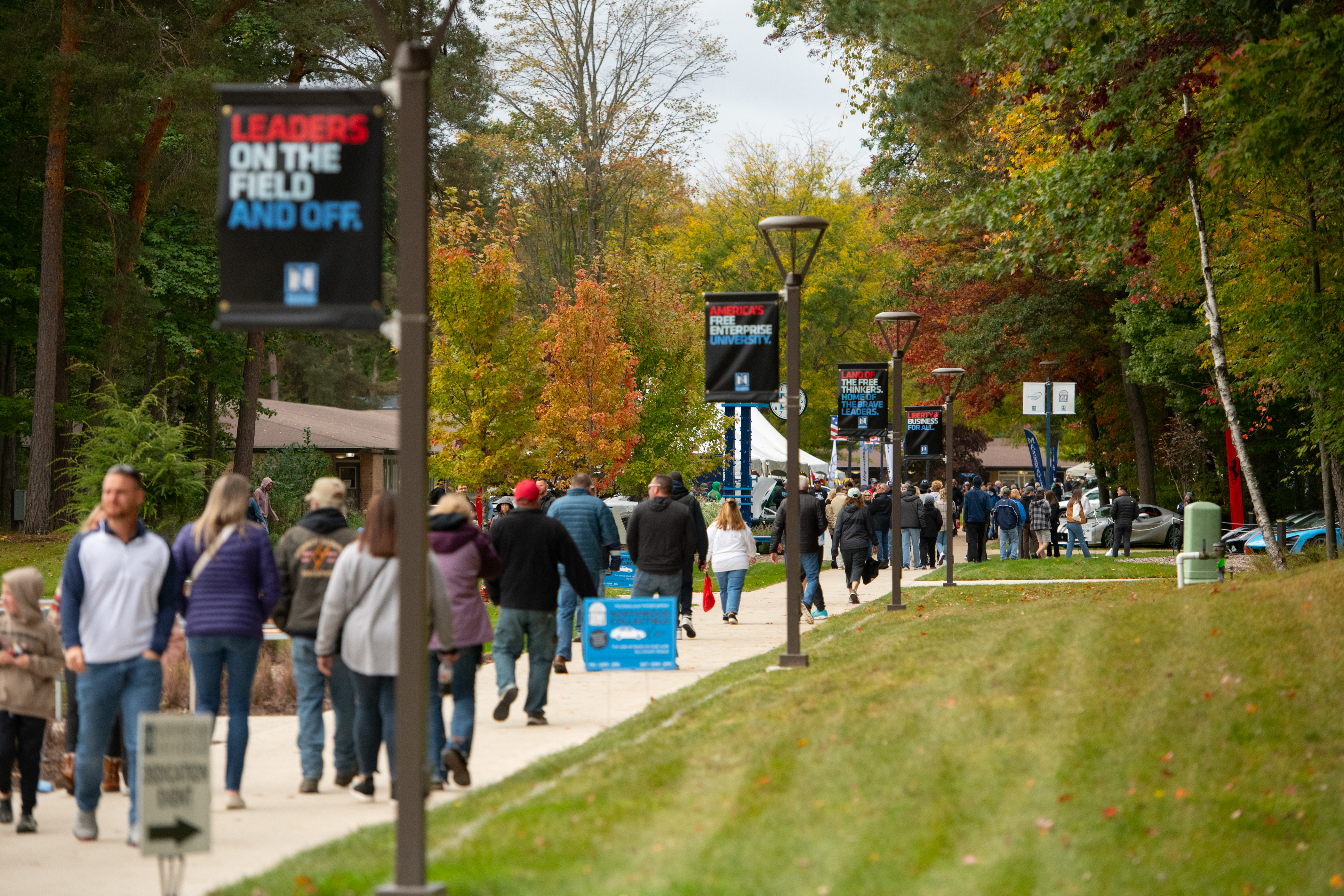 People walk along a campus pathway lined with trees and lamp posts displaying banners that read "Leaders on the field and off," during the Annual Alumni Gathering. It appears to be autumn.