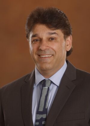 A man in a dark suit and patterned tie smiles at the camera against a plain brown background.