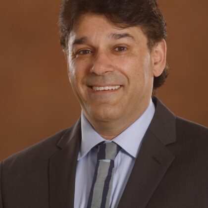 A man in a dark suit and patterned tie smiles at the camera against a plain brown background.
