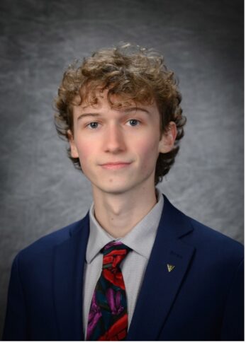 A young man with curly hair wearing a blue suit, patterned tie, and light-colored shirt poses for a studio portrait against a gray backdrop.