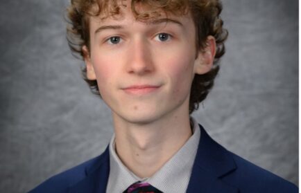 A young man with curly hair wearing a blue suit, patterned tie, and light-colored shirt poses for a studio portrait against a gray backdrop.