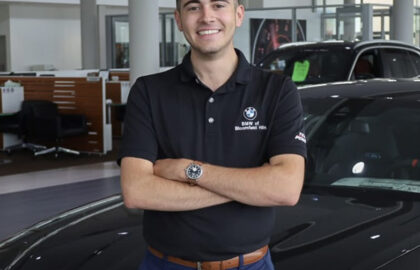 A man in a black BMW polo shirt stands with arms crossed in front of a black car inside Motor City’s modern car dealership showroom.