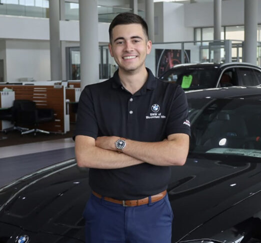 A man in a black BMW polo shirt stands with arms crossed in front of a black car inside Motor City’s modern car dealership showroom.
