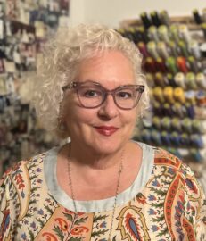 A woman with curly white hair, glasses, and a patterned top smiles at the camera. Behind her are spools of thread and photos pinned to a wall, celebrating her recognition in Northwood's Distinguished Women Awards.