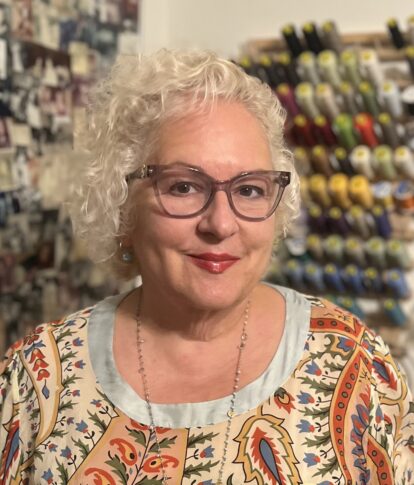 A woman with curly white hair, glasses, and a patterned top smiles at the camera. Behind her are spools of thread and photos pinned to a wall, celebrating her recognition in Northwood's Distinguished Women Awards.