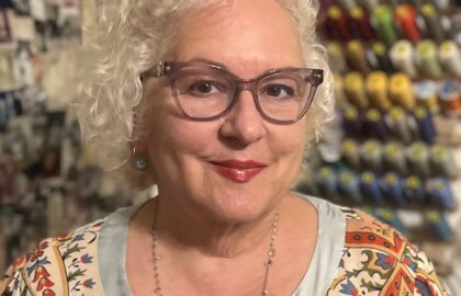 A woman with curly white hair, glasses, and a patterned top smiles at the camera. Behind her are spools of thread and photos pinned to a wall, celebrating her recognition in Northwood's Distinguished Women Awards.