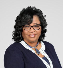 Woman with black curly hair wearing glasses, a navy blazer with white trim, and a blue beaded necklace, posing against a plain light background for the Northwood's Distinguished Women Awards.