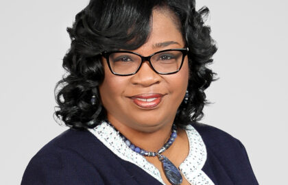 Woman with black curly hair wearing glasses, a navy blazer with white trim, and a blue beaded necklace, posing against a plain light background for the Northwood's Distinguished Women Awards.