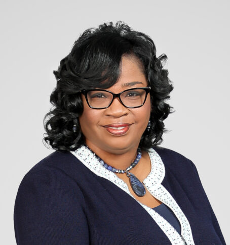 Woman with black curly hair wearing glasses, a navy blazer with white trim, and a blue beaded necklace, posing against a plain light background for the Northwood's Distinguished Women Awards.
