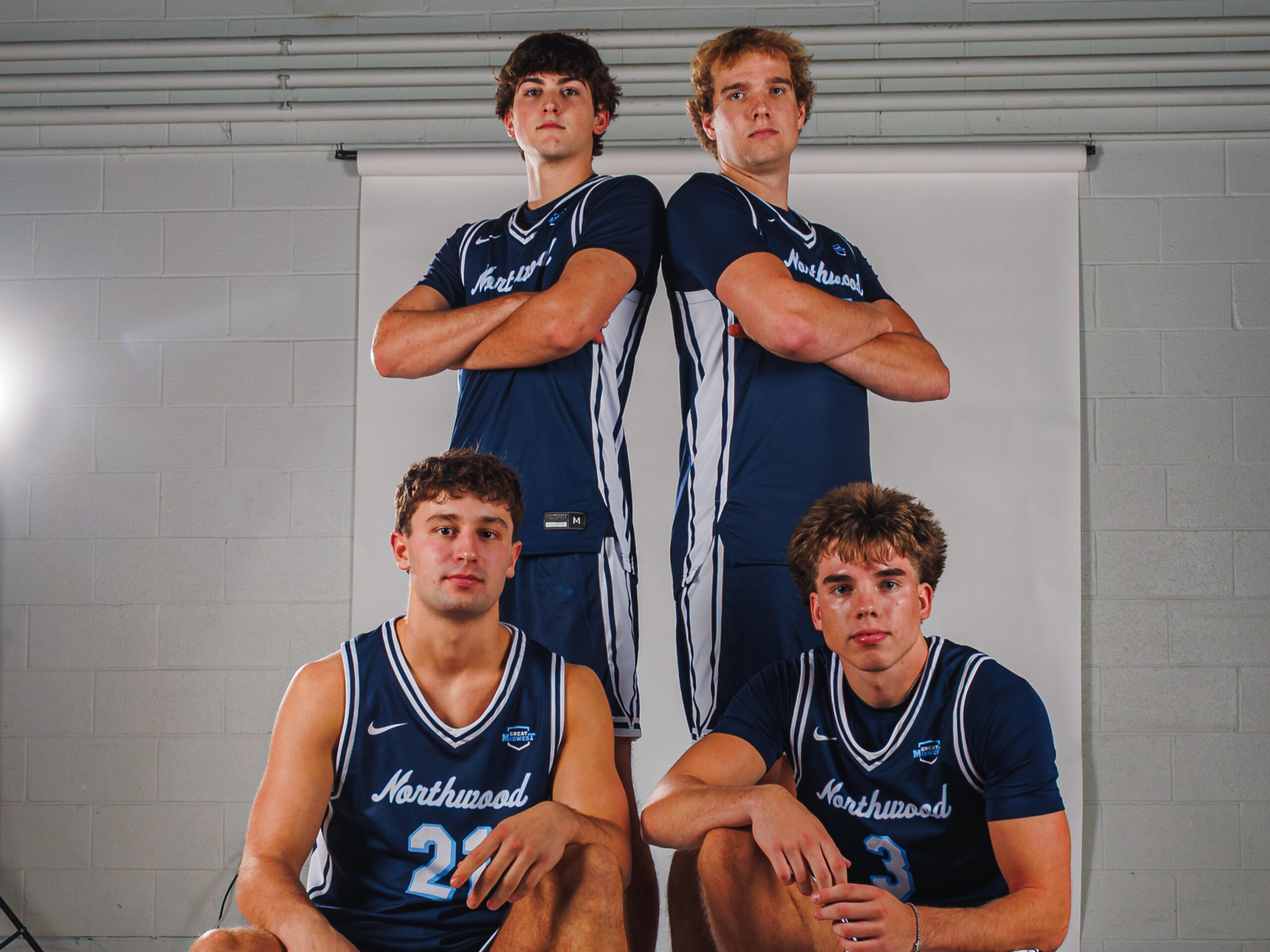 Four male basketball players in blue Northwood uniforms pose in a gym, two standing with arms crossed and two seated on basketballs, representing the Sixth Man Club, in front of a white backdrop.