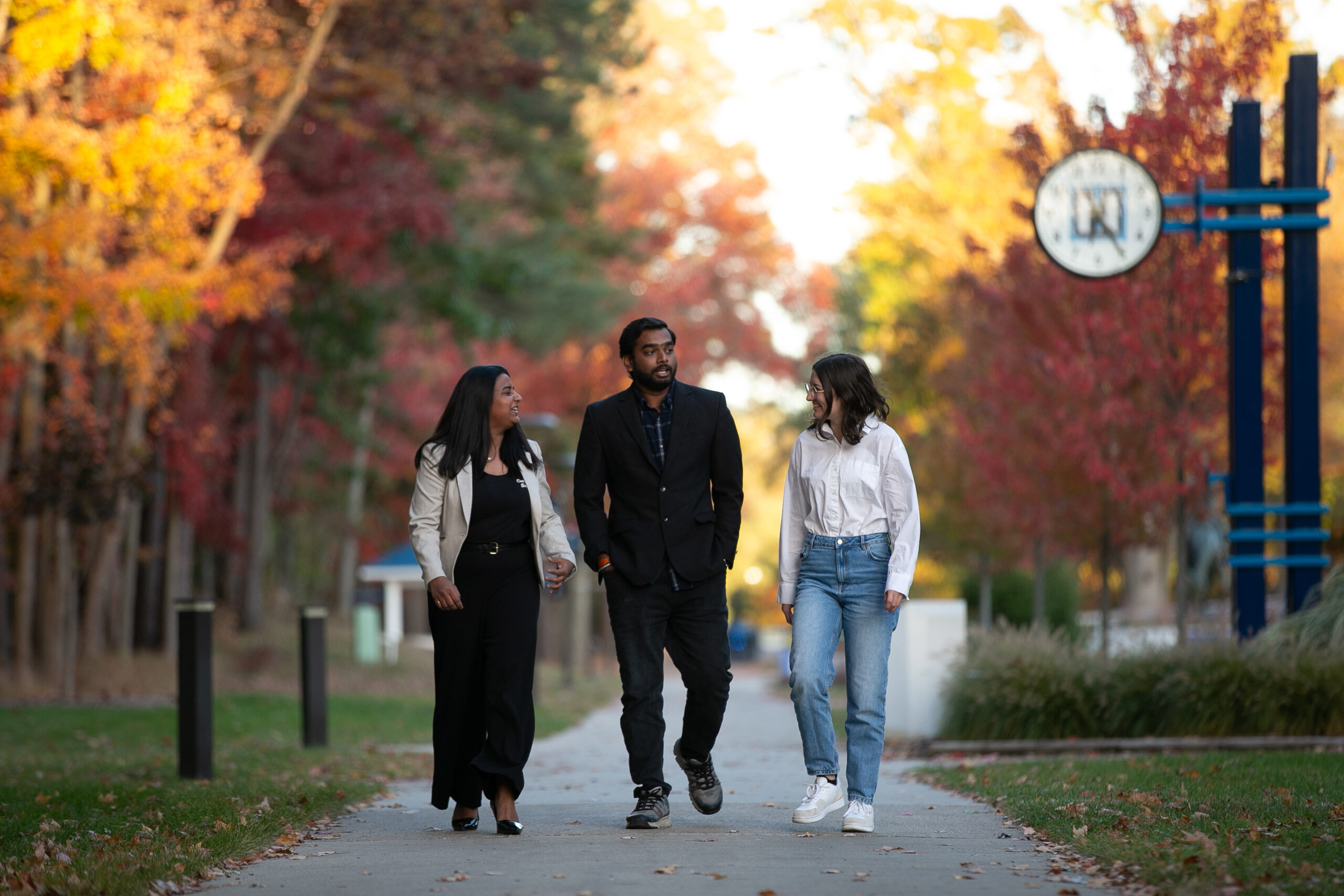 Three people walk and talk on a tree-lined path in autumn, colorful foliage surrounding them and a Timberwolf Tracks sign visible in the blue and white background.