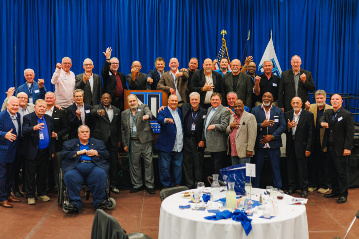 A large group of men in suits and business attire pose for a group photo in front of blue curtains, with flags and a podium in the background at an indoor event.