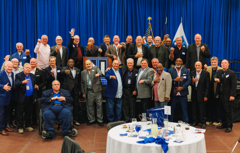 A large group of men in suits and business attire pose for a group photo in front of blue curtains, with flags and a podium in the background at an indoor event.
